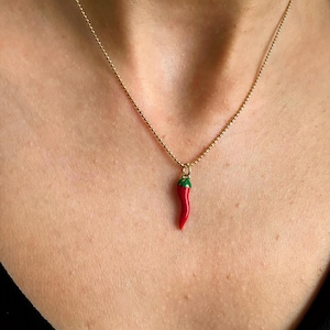 May include: A gold chain necklace featuring a small, red chilli pepper pendant with green detailing. The pendant is a bright red colour and is suspended from a fine chain. The necklace is worn around the neck.
