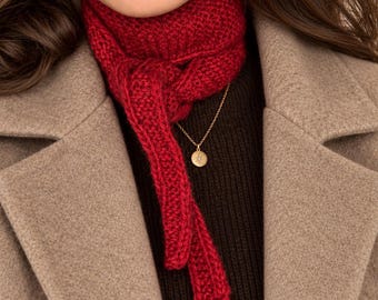 May include: A deep red knitted scarf tied in a simple knot around the neck, worn over a dark brown ribbed sweater and a beige wool coat. A delicate gold chain necklace with a small round pendant featuring a star design rests against the sweater.