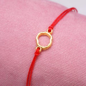 May include: A red string bracelet with a gold circle charm. The charm has an irregular, organic shape.