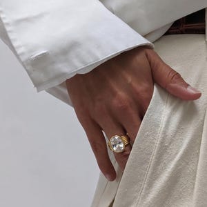 Puede incluir: Un anillo de oro con una gran gema clara de corte ovalado. El anillo tiene un diseño acanalado en la banda y está sobre un fondo blanco. El anillo es una pieza llamativa.