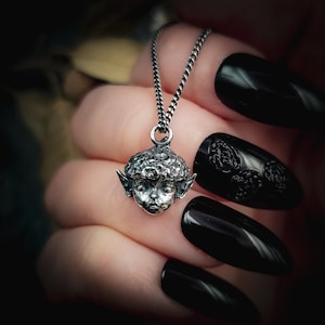 May include: A silver pendant necklace featuring a detailed, stylized elf face with pointed ears and textured hair. The pendant hangs from a delicate chain. The elf's face is intricately detailed, and the necklace is shown against a blurred background.