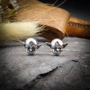 May include: A pair of silver earrings featuring a detailed face with pointed ears. The earrings are small and delicate, perfect for adding a touch of whimsy to any outfit.