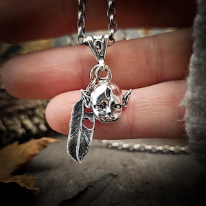 May include: A silver necklace with a pendant featuring a small, detailed sculpture of a creature with a feather. The creature has a pointed head with a third eye and large ears.