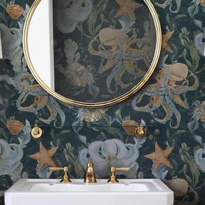 Op de afbeelding: Badkamerinterieur met een witte wastafel, gouden armaturen en een ronde, goudomrande spiegel. Het behang toont een onderwaterlandschap met octopussen, zeesterren en schelpen. Twee gouden wandlampen met witte kappen.