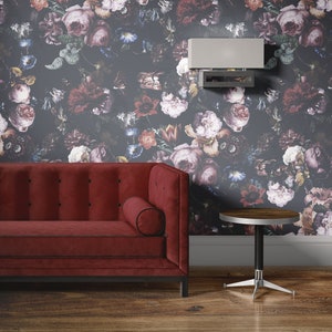 May include: A red velvet sofa with a black trim and button tufting sits in front of a dark floral wallpaper. A small round wooden table with a silver base sits to the right of the sofa. A silver light fixture is mounted on the wall above the sofa.