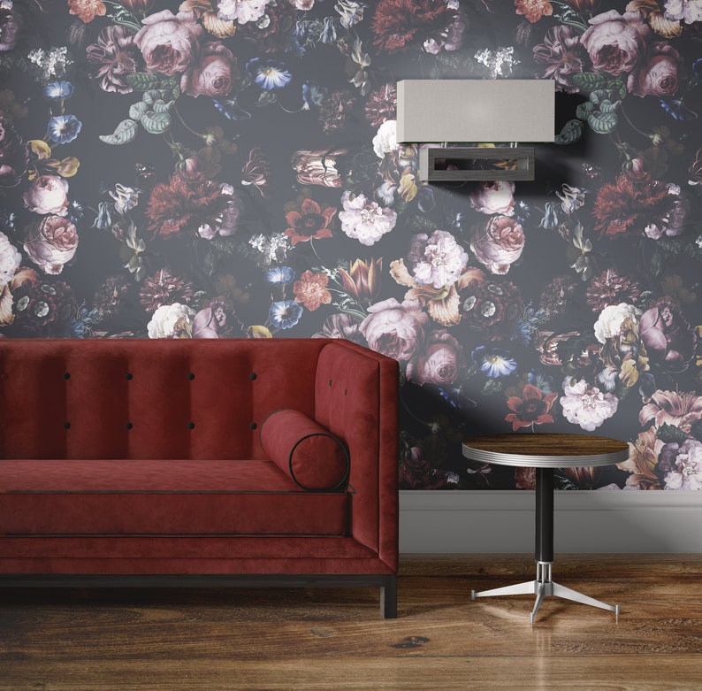 May include: A red velvet sofa with a black trim and button tufting sits in front of a dark floral wallpaper. A small round wooden table with a silver base sits to the right of the sofa. A silver light fixture is mounted on the wall above the sofa.