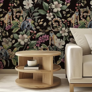 May include: A light wood, two-tiered round coffee table with a small bowl and books on the middle shelf. The background features a black floral wallpaper with monkeys, flowers, and snakes. A beige sofa is on the right.