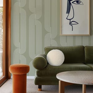 Könnte beinhalten: Ein Wohnzimmer mit einem grünen Samtsofa, einem orangefarbenen Samthocker und einem runden Couchtisch. Die Wand ist mit einem gerahmten abstrakten Gesichtskunstwerk und einer salbeigrünen Tapete mit weißen vertikalen Linien geschmückt. Ein flauschiges weißes rundes Kissen liegt auf dem Sofa.