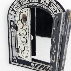 Peut inclure: Miroir mural décoratif noir et blanc, avec un sommet en forme d'arche et une porte à charnière. Le miroir présente des motifs et des dessins détaillés, notamment des motifs floraux et en volutes. La porte est entrouverte, révélant la surface miroir.