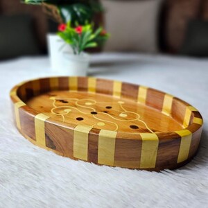 Peut inclure: Un plateau ovale en bois avec incrustation décorative. Le plateau présente des rayures alternées de bois clair et foncé autour du bord. L'intérieur a une incrustation en bois clair avec un motif tourbillonnant et de petits découpes circulaires.
