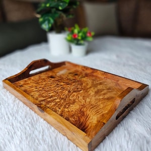 Può includere: Un vassoio rettangolare in legno con un motivo a venature naturali. Il vassoio ha maniglie integrate su ogni lato ed è realizzato in legno marrone chiaro. Il vassoio è appoggiato su una superficie bianca e soffice.