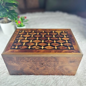 Peut inclure: Une boîte rectangulaire en bois avec une finition en loupe. Le couvercle présente un motif géométrique incrusté complexe composé de losanges marron foncé et beige clair. Une petite serrure est centrée sur la face avant de la boîte.