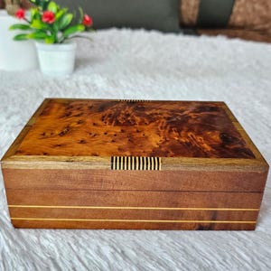 Peut inclure: Une boîte rectangulaire en bois dotée d'un couvercle en loupe de bois brun foncé riche avec un motif de grain naturel tourbillonnant. La boîte est accentuée par de fines rayures incrustées horizontales claires et des détails décoratifs rayés noir et blanc.