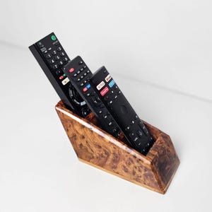 May include: A wooden remote control holder with a burl wood grain pattern. Three black remote controls are inside the holder. The holder is angled, with the remotes partially visible.
