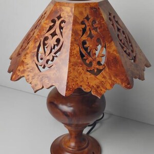 May include: A wooden table lamp with a carved shade. The shade is a light brown color with intricate cut-out designs. The base is a darker brown, with a round bulbous shape and a pedestal base. The lamp is plugged in.