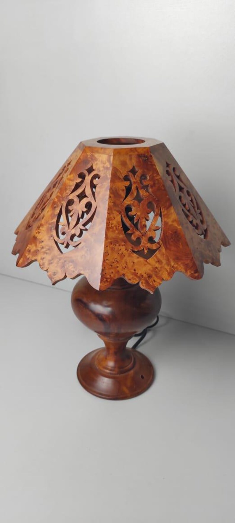 May include: A wooden table lamp with a carved shade. The shade is a light brown color with intricate cut-out designs. The base is a darker brown, with a round bulbous shape and a pedestal base. The lamp is plugged in.