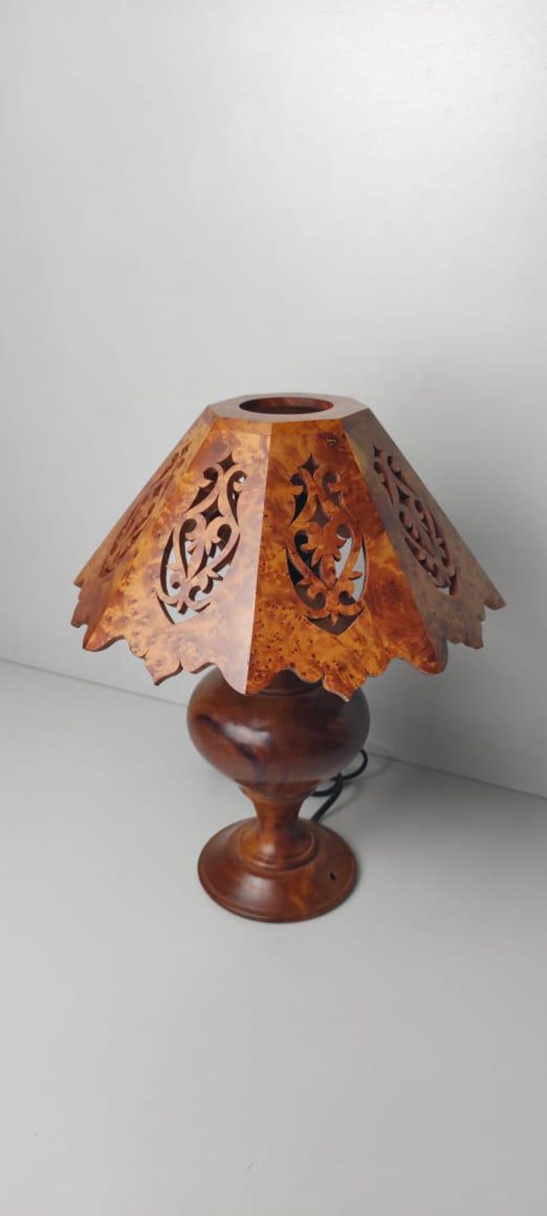 May include: A wooden table lamp with a decorative shade. The shade is a light brown color with intricate cut-out designs. The base is a darker brown, with a round bulbous shape and a pedestal base.
