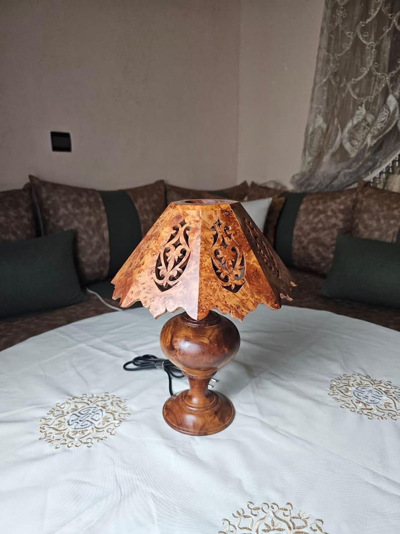 May include: A wooden table lamp with a carved, patterned shade and a round base. The shade is a light brown color with intricate cut-out designs. The base and stem are a darker brown, and the lamp is on a white tablecloth.
