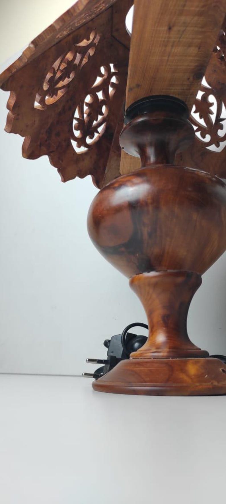May include: A wooden table lamp with a carved shade featuring intricate floral cutouts. The lamp has a bulbous base and a dark brown finish, with a power cord and plug visible.