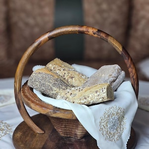 Op de afbeelding: Een houten broodmand met een gebogen handvat, gevuld met diverse broden. De mand staat op een witte doek met een geborduurd ontwerp. Het brood heeft een verscheidenheid aan zaden en granen.