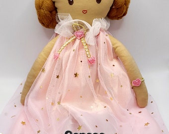 May include: A handmade doll with brown hair, wearing a pink tulle dress with gold stars and a pink heart-shaped bracelet. The doll is sitting on a white background. The text "Genese" is visible on the dress.