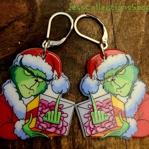 May include: A pair of Grinch-themed Christmas earrings. Each earring features a green Grinch wearing a Santa hat and holding a box of pink hearts. The earrings are made of acrylic and have silver hooks.