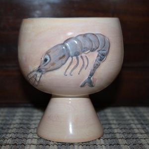 May include: A ceramic goblet with a hand-painted design of a shrimp. The goblet has a light pink base and a white rim.