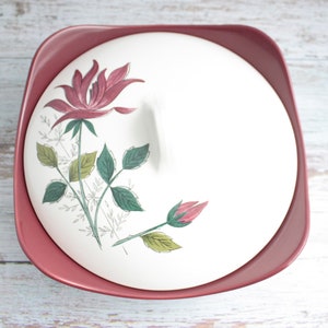 May include: A vintage serving dish with a lid, featuring a floral design. The dish is a dusty rose colour, with a white lid adorned with a pink rose and green leaves. The square dish is likely made of ceramic.