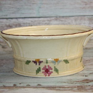 May include: A cream-coloured ceramic bowl with two handles and a floral design. The bowl has a brown rim and a floral pattern with green leaves, purple, yellow, and blue flowers, and a pink flower in the centre.
