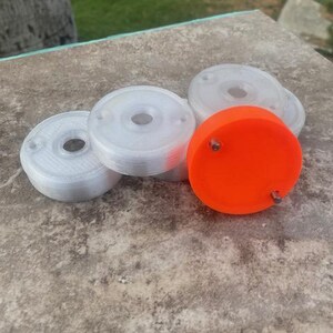 May include: A collection of circular, disc-shaped components featuring a central hole and two small metal pins protruding from the surface. Several discs are translucent white, while one prominent disc is a vibrant, solid neon orange color.