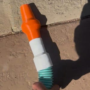 May include: An orange and white garden hose nozzle attached to a teal corrugated hose. The nozzle is held by a hand, with the hose resting on a textured, light brown surface. The image is taken outdoors.
