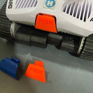 May include: A white and black robotic pool cleaner with an orange "PUSH" button and a blue component. The cleaner has black wheels and the brand name "HAYWARD" is visible. The cleaner has a blue and white logo.