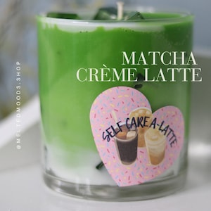 May include: A green and white candle in a glass jar with the text "MATCHA CREME LATTE" on the top and a pink heart sticker with the text "SELF CARE A-LATTE" on the front.