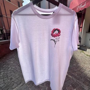 Può includere: T-shirt bianca con girocollo e maniche corte. La maglietta presenta un disegno floreale rosso e nero con un volto e le parole "Bella Ciao" in grafia nera. La maglietta è appesa a una gruccia nera.