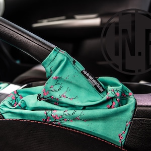 Peut inclure: Un soufflet de levier de vitesse de voiture à motif turquoise et rose avec le texte "easy the clutch, shift the fun" imprimé sur le côté.