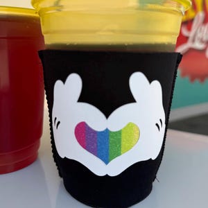 May include: A black neoprene cup sleeve featuring a white graphic of two cartoon gloved hands forming a heart shape. The center of the heart is filled with a vertical rainbow-colored pattern. The sleeve is wrapped around a clear plastic cup.