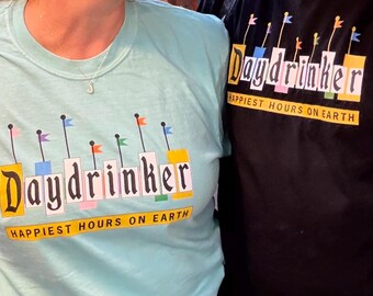 May include: Two people wearing matching t-shirts. One shirt is light blue, the other is black. Both shirts have the text "Daydrinker" in a stylized font, with colorful flag accents and the phrase "Happiest Hours On Earth" below.
