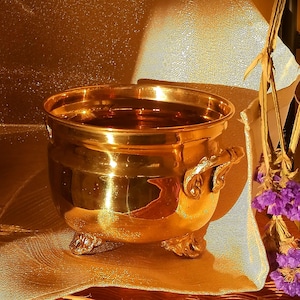 May include: A gold-colored metal pot with three legs and a handle. The pot is sitting on a gold-colored fabric surface.