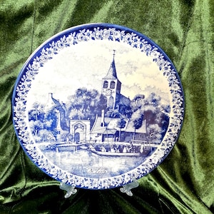Peut inclure: Une assiette en céramique bleue et blanche représentant une scène de village hollandais. L'assiette présente une église, des maisons, des arbres et un canal. L'assiette est décorée d'une bordure florale et du texte "De Lente" au centre.