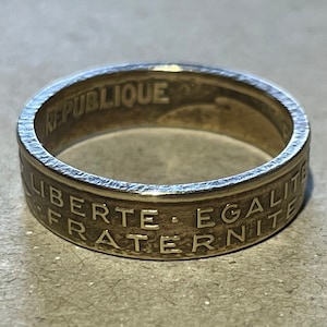 May include: A gold-colored ring made from a French coin. The ring features the inscription "REPUBLIQUE" on the top and "LIBERTE - EGALITE - FRATERNITE" on the bottom.
