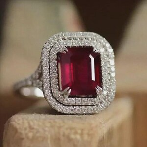 May include: A silver-toned ring featuring a large, rectangular, deep red gemstone at the center. The stone is surrounded by a double halo of small, clear, sparkling stones. The band is split and also encrusted with small, clear, sparkling stones.