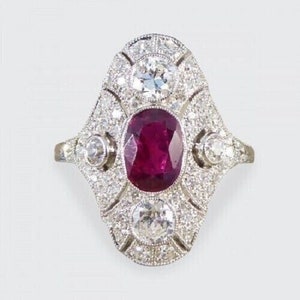 Könnte beinhalten: Ein silberner Ring mit einem großen ovalen Rubin, der von kleineren Diamanten umgeben ist. Der Ring hat ein zartes, filigranes Design.