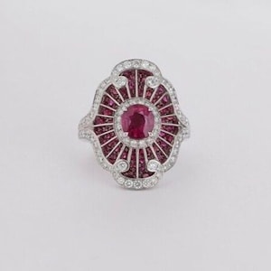 May include: A silver ring with a large oval ruby surrounded by smaller rubies and diamonds. The ring has a decorative design with a fan-like pattern.