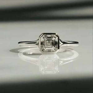 May include: A silver ring featuring a square-cut diamond. The diamond is set within a bezel, and the band is slender. The ring is shown on a reflective surface, creating a mirrored effect.