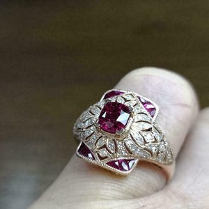 Peut inclure: Une bague en or avec un gros rubis de taille carrée entouré de diamants plus petits. La bague a un design délicat et complexe avec un motif floral.