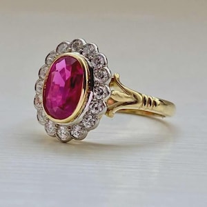 May include: A gold ring featuring a central oval-cut vibrant pink gemstone set in a yellow gold bezel. The center stone is surrounded by a halo of small round white diamonds set in white metal, with a polished yellow gold band and decorative shoulders.