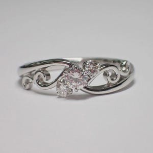 Könnte beinhalten: Ein silberner Ring mit drei klaren, runden Edelsteinen. Der Ring hat ein zartes, wirbelndes Design auf beiden Seiten der Edelsteine, was einen eleganten und zeitlosen Look erzeugt. Der Ring ist vor einem schlichten weißen Hintergrund platziert.