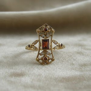 May include: A gold ring featuring an elongated, openwork metal design with a central rectangular garnet gemstone. The setting is accented with smaller round garnet stones and tiny clear stones, arranged in a symmetrical, intricate pattern.