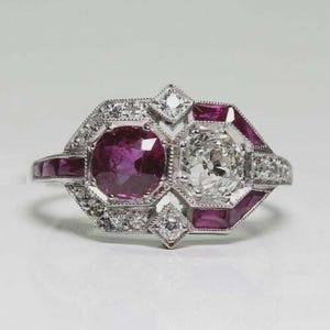 May include: A vintage-style ring featuring a large, round, deep-purple ruby and a clear, sparkling diamond. The ring is set in a silver or white gold band, with smaller diamonds and ruby accents. The design is geometric and elegant.