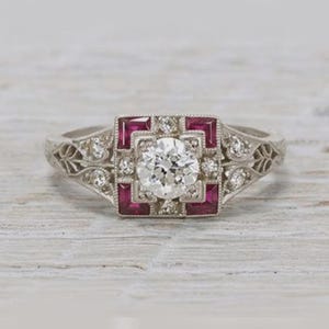 Peut inclure: Bague de style vintage avec une grosse pierre centrale ronde et transparente, entourée de petites pierres transparentes et de pierres rouges rectangulaires. Le corps de la bague est argenté avec des motifs floraux et de feuilles.